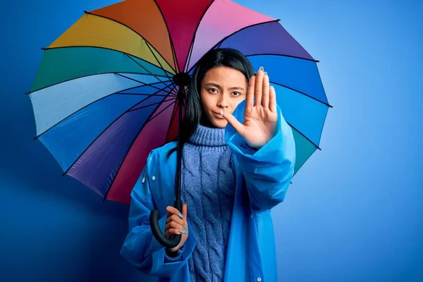 年轻的中国女人穿着雨衣 拿着雨伞遮挡在孤零零的蓝色背景上 张开双手 做了一个严肃而自信的手势 做了一个防守姿势 — 图库照片