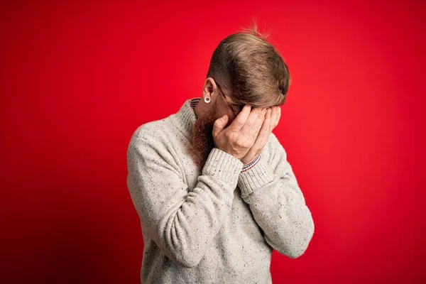 Handsome Irish redhead man with beard wearing casual sweater and glasses over red background with sad expression covering face with hands while crying. Depression concept.