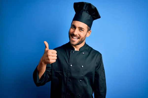 ひげを生やした若いハンサムなシェフの男は 炊飯器の制服を着て 青い背景の上に帽子を手で幸せな親指を立ててジェスチャーをします 成功を示すカメラを見て表現を承認する — ストック写真