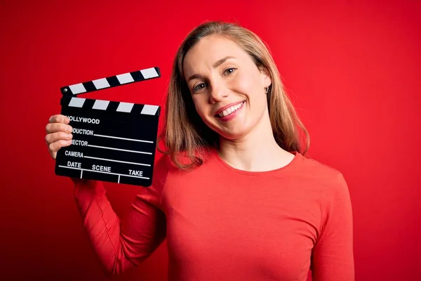 Joven Hermosa Rubia Aplaudiendo Mujer Sosteniendo Clapboard Sobre Fondo Rojo —  Fotos de Stock