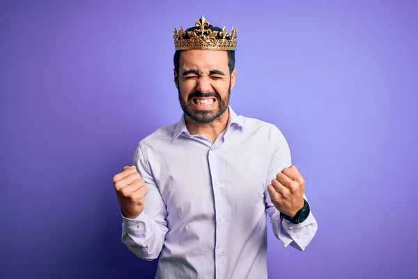 年轻英俊的男人留着胡子 头戴金色的王冠 背景是紫色的 他非常高兴而兴奋地举起双臂 微笑着 尖叫着要成功 庆祝概念 — 图库照片