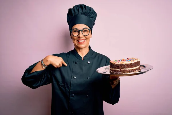 中年妇女烘焙师 身穿炊具制服 头戴礼帽 端着美味的蛋糕 满脸惊讶 手指指向自己 — 图库照片