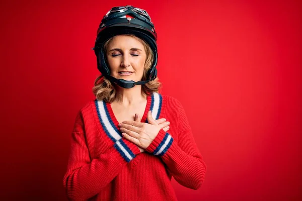 中世の美しいブロンドのオートバイの女性は 目を閉じて胸に手で笑みを浮かべて赤い背景に元ヘルメットを身に着けており 顔に感謝のジェスチャー 健康コンセプト — ストック写真