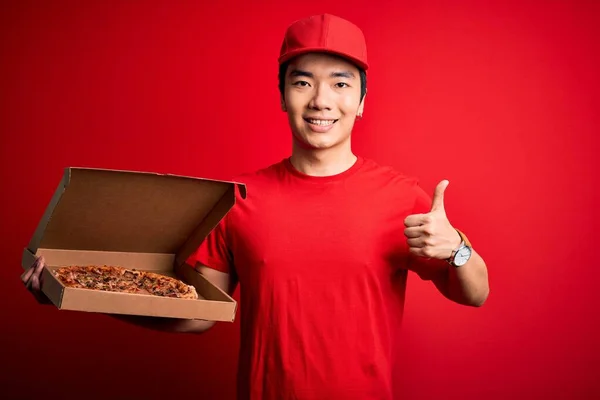 Young handsome chinese delivery man holding deliver box with delicious Italian pizza happy with big smile doing ok sign, thumb up with fingers, excellent sign