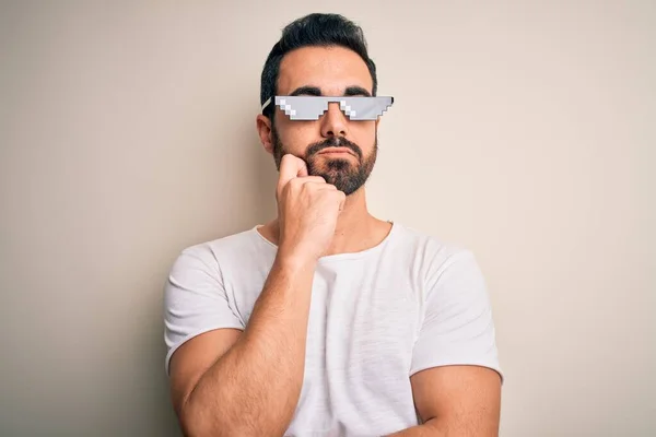 Young handsome man with beard wearing funny thug life sunglasses over white background with hand on chin thinking about question, pensive expression. Smiling with thoughtful face. Doubt concept.
