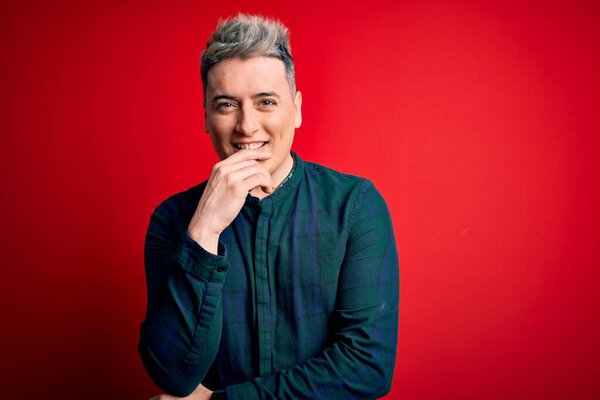 Young handsome modern man wearing elegant green shirt over red isolated background looking confident at the camera smiling with crossed arms and hand raised on chin. Thinking positive.