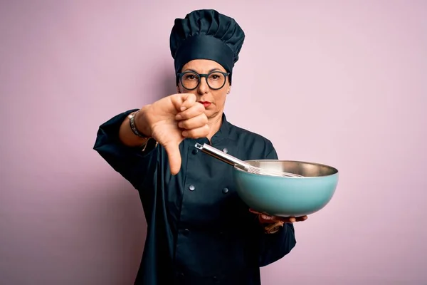 Middle age brunette chef woman wearing cooker uniform and hat using whisk and bowl with angry face, negative sign showing dislike with thumbs down, rejection concept