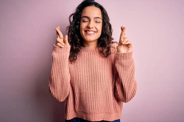 Young beautiful woman with curly hair wearing casual sweater over isolated pink background gesturing finger crossed smiling with hope and eyes closed. Luck and superstitious concept.