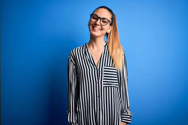Beautiful blonde woman with blue eyes wearing striped shirt and glasses over blue background with a happy and cool smile on face. Lucky person.