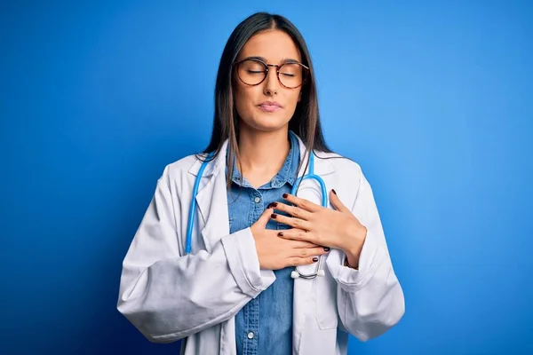 Young beautiful doctor woman wearing stethoscope and glasses over blue background smiling with hands on chest with closed eyes and grateful gesture on face. Health concept.