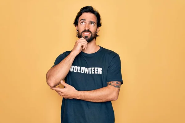 若いハンサムなヒスパニック系のボランティアの男性の質問について考えてあごの手で社会的ケアとしてボランティアTシャツを着て 包括的な表現 笑顔と思慮深い顔 疑いの概念 — ストック写真