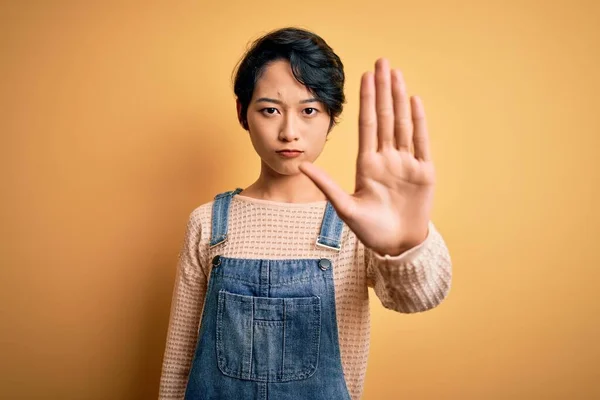 年轻美丽的亚洲女孩穿着休闲装 在孤立的黄色背景上做着停止手心歌唱的工作 脸上带有消极而严肃的手势的警告表情 — 图库照片