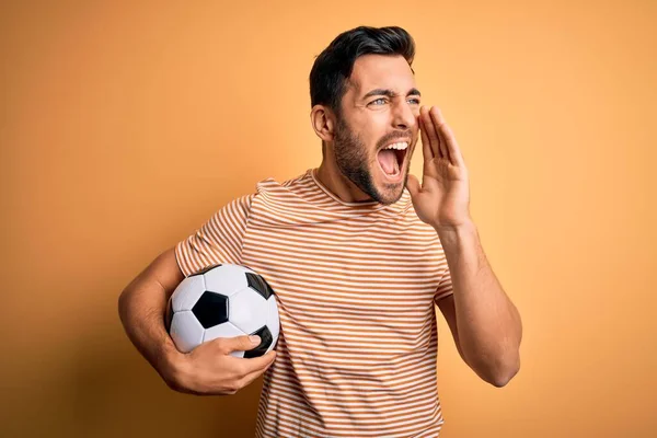 黄色い背景の叫びながらサッカーボールを持っている髭を生やしたハンサムなプレーヤーの男と大声で手を口の中に横に叫んでいる コミュニケーションの概念 — ストック写真