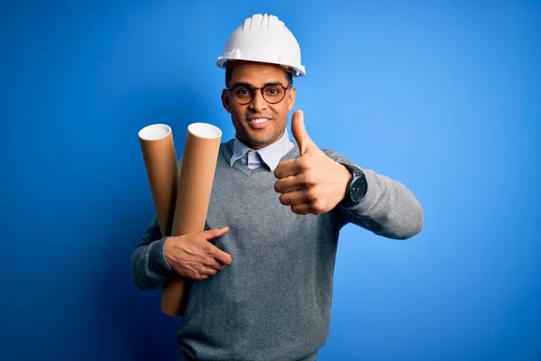 Young african american afro architect man wearing safety helmet holding bluprints happy with big smile doing ok sign, thumb up with fingers, excellent sign