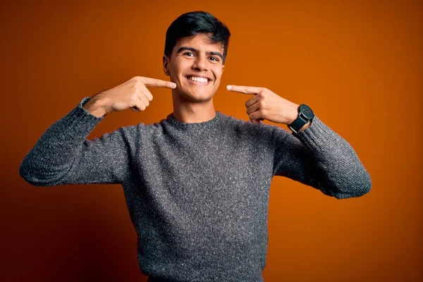 İzole edilmiş turuncu arka planda dikilen sıradan bir süveter giyen genç, neşeli bir şekilde gülümseyen ve parmaklarıyla ve ağzıyla işaret eden bir adam. Diş sağlığı konsepti.