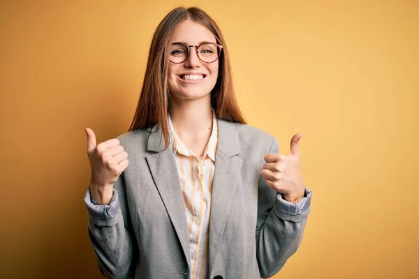 年轻美丽的红头发女人 穿着夹克和眼镜 戴着孤立的黄色背景的成功标志 手拿着积极的手势 微笑着竖起大拇指 快乐地做着 快乐的表情和胜利的姿态 — 图库照片