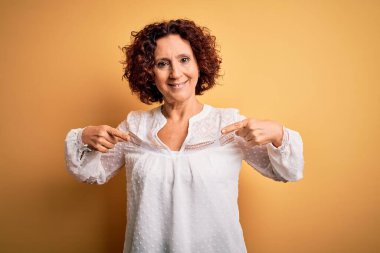 Orta yaşlı güzel kıvırcık saçlı kadın sarı arka planda rahat bir yaz elbisesi giyiyor. Kendine güler yüzlü, gururlu ve mutlu birini işaret ediyor..