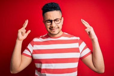 İzole edilmiş kırmızı arka plan üzerinde sıradan çizgili tişört ve gözlük takan genç yakışıklı adam umut ve gözler kapalı bir şekilde gülümseyerek işaret ediyor. Şans ve batıl inanç..