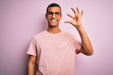 Yakışıklı Afro-Amerikalı adam pembe arka planda günlük tişört ve gözlük takıyor gülümsüyor ve el hareketleriyle el hareketleriyle küçük boy işareti yapıyor ve kameraya bakıyor. Ölçü kavramı.