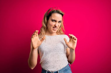 Genç, sarışın, günlük tişört giyen, pembe arka planda duran, tiksindirici, tiksindirici, tiksindirici bir yüz ifadesi takınan genç bir kadın. Eller havada