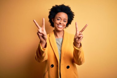 Kıvırcık saçlı, sarı ceketli, gülümseyen ve parmaklarını zafer işareti yaparken gösteren Afro-Amerikan genç, güzel iş kadını. İki numara..