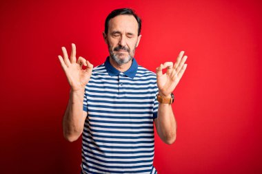 İzole edilmiş kırmızı arka planda dikilen sıradan çizgili polo giyen orta yaşlı adam rahatla ve parmaklarıyla meditasyon hareketi yaparken gözleri kapalı gülümse. Yoga konsepti.