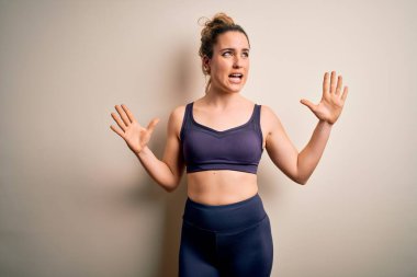 Beyaz arka plan üzerine spor giyerek spor yapan genç ve güzel sarışın kadın. Deli gibi bağırıyor, bağırıyor, agresif ifadeler ve kollar havada. Hayal kırıklığı konsepti.