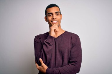 Beyaz arka plan üzerine günlük süveter giyen genç ve yakışıklı Afro-Amerikan adam kameraya kollarını kavuşturup gülümseyerek bakıyor ve çenesini kaldırıyor. Olumlu düşünüyorum..