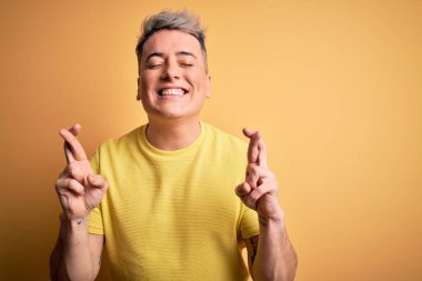 Sarı izole edilmiş arka plan üzerine sarı tişört giyen genç ve modern adam umut ve gözler kapalı bir şekilde gülümseyerek işaret ediyor. Şans ve batıl inanç..