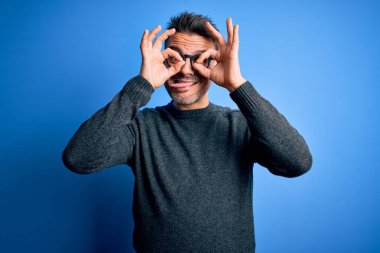 Mavi arka planda duran sıradan süveter ve gözlük takan genç yakışıklı bir adam dürbün gibi dil çıkartıyor, gözler parmaklarından bakıyor. Çılgın ifade.