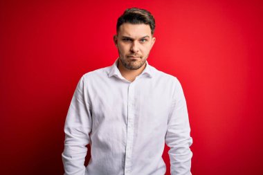 Mavi gözlü, zarif gömlekli genç bir iş adamı kırmızı izole edilmiş arka planda duruyor şüpheci ve gergin, sorun yüzünden suratını asıyor. Negatif kişi.