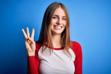 Genç, güzel, kızıl saçlı bir kadın, mavi arka planda rahat bir tişört giyip üç numaralı parmaklarıyla işaret ederken, kendinden emin ve mutlu bir şekilde gülümsüyor..