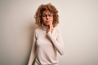 Balıkçı yaka kazak giyen ve beyaz arka plan üzerine gözlük takan genç Afro-Amerikan bir kadın diş ağrısı ya da diş ağrısı yüzünden ağız ve acı dolu bir ifadeyle dokunuyor. Dişçi