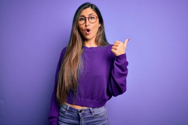 Mor izole edilmiş arka planda gözlük takan genç, güzel, zeki bir kadın. El parmağı yana dönük, ağzı açık, şaşkın bir ifade..
