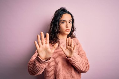 Kıvırcık saçlı, pembe arka planda günlük süveter giyen genç ve güzel bir kadın. Avuçlarını hareket ettiriyor, reddediyor ve iğrenç bir ifadeyle reddediyor. Dur ve yasakla.