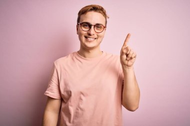 İzole edilmiş pembe arka planda duran, gündelik tişört giyen genç, kızıl saçlı adam başarılı bir fikir işaret ediyor. Heyecanlı ve mutlu. Bir numara..