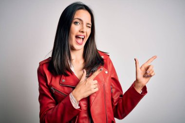 Mavi gözlü genç esmer kadın beyaz arka planda kırmızı bir ceket giyiyor gülümsüyor ve kameraya bakıyor iki eliyle ve parmaklarıyla yandan işaret ediyor..