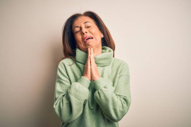 Orta yaşlı, güzel bir kadın, izole edilmiş beyaz arka plan üzerine balıkçı yaka kazak giyip elleri bağlı yalvarıyor ve dua ediyor. Yüzünde umut ifadesi var. Çok duygusal ve endişeli. Yalvarıyorum..