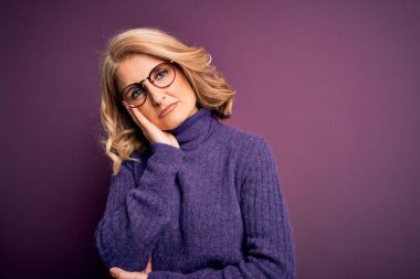 Orta yaşlı, sarışın, sıradan mor balıkçı yaka kazak ve gözlük giyen güzel bir kadın kollarını kavuşturarak depresyondan sıkılmış ve yorgun göründüğünü düşünüyor..