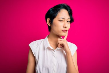 Genç ve güzel Asyalı bir kız günlük yaz gömleği giyiyor pembe arka planda duruyor, elleri çenesinde soru sorarken, düşünceli bir yüz ifadesi var. Düşünceli bir yüzle gülümsüyor. Şüphe kavramı.