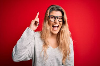 İzole edilmiş kırmızı arka plan üzerinde süveter ve gözlük takan genç, güzel sarışın kadın başarılı bir fikri işaret ediyor. Heyecanlı ve mutlu. Bir numara..
