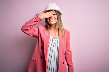 Genç ve güzel esmer mimar kadın pembe arka planda güvenlik kaskı takıyor gülümsüyor ve elleri yüzünde şaşkınlıktan gözleri kapayarak gülüyor. Kör kavram.