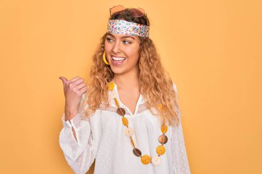 Genç, sarışın, mavi gözlü, güneş gözlüklü, aksesuar takan, gülümseyen, gülümseyen ve baş parmağıyla yüzünü gösteren güzel bir hippi kadın..