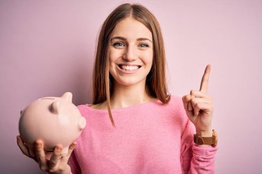 Genç, kızıl saçlı, kumbarayı soyutlanmış pembe arka planda tutan güzel bir kadın bir fikir ya da soru işareti ile şaşırdı.