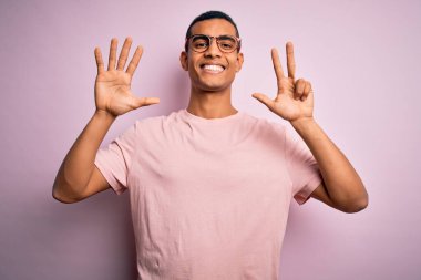 Yakışıklı Afro-Amerikalı adam pembe arka planda günlük tişört ve gözlük takıyor ve parmaklarını 8 numaraya doğrultuyor..