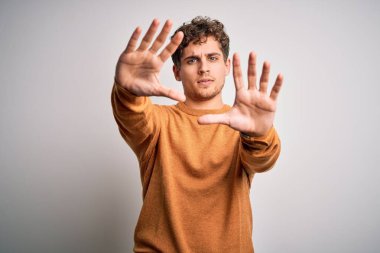 Sarışın, kıvırcık saçlı, beyaz arka plan üzerine günlük süveter giyen, elleri ve parmakları kullanarak çerçeve yapan, kamera perspektifli genç, yakışıklı bir adam.