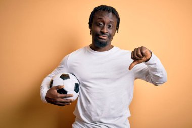 Genç Afrikalı Amerikalı oyuncu futbol topunu sarı zemin üzerinde tutarak oynuyor. Kızgın yüzlü, negatif işareti var. Başparmakları aşağıda, reddedilme konsepti.