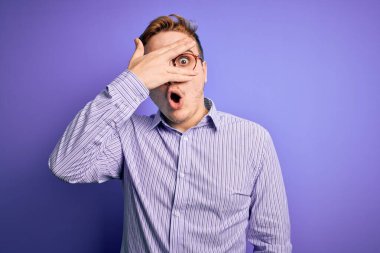 Genç, kızıl saçlı, sıradan bir gömlek ve mor arka planda gözlük takmış. Şok olmuş bir şekilde yüzünü ve gözlerini kapayarak utanmış bir ifadeyle parmaklarının arasından bakıyor..