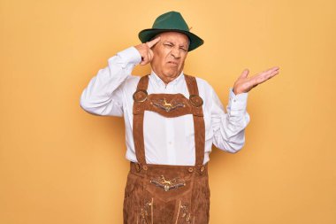 Sarı arka planda Alman geleneksel ahşap şenliği kıyafeti giyen yaşlı gri saçlı bir adam. Kafası karışmış ve avuç içi açık bir şekilde fotokopi alanı göstermekten ve alnını işaret etmekten rahatsız olmuş. Bunu bir düşün..
