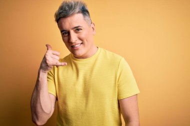 Sarı izole edilmiş arka plan üzerinde sarı tişörtlü, genç ve modern bir adam telefonla el ve parmak hareketleriyle gülümsüyor, telefonda konuşuyor gibi. İletişim kavramları.